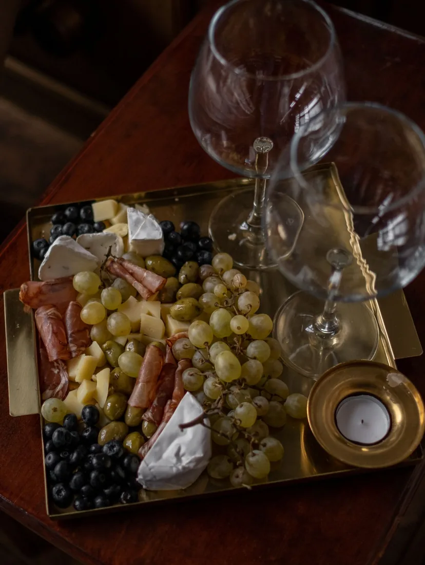Seen from above, a brass tray of cured meat, soft cheeses, green grapes and olives beside two empty wine glasses