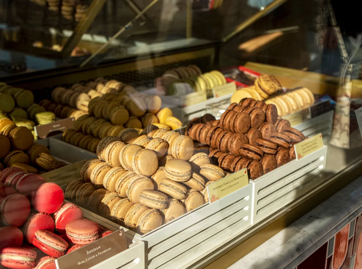 Trays of pale and chocolate-brown macarons stacked behind a glass shopfront counter, with pink ones in the corner