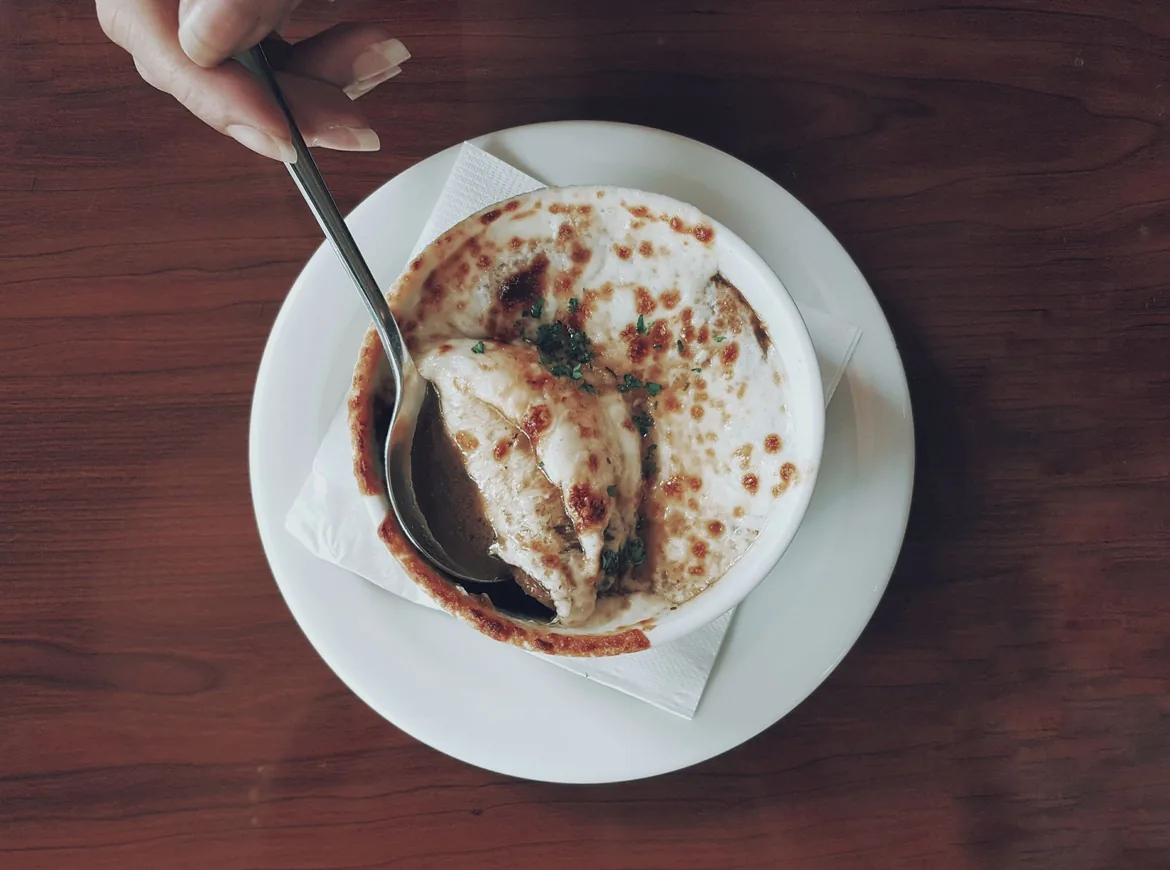 Seen from above, a spoon lifting the browned cheese crust of French onion soup in a white bowl